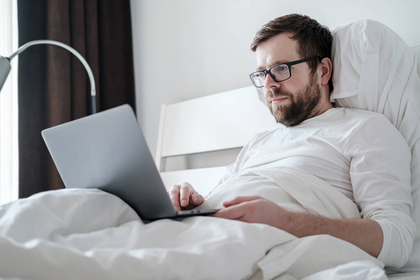 Wolny strzelec. Zadowolony, młody mężczyzna w piżamie pracuje z laptopem, leżąc w miękkim łóżku, w komfortowych apartamentach. - Zdjęcie, obraz