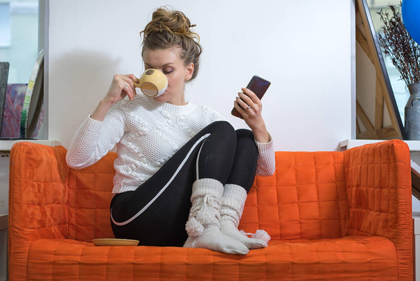 Kadın akıllı telefonuyla kanepeye oturur ve kahve içer. - Fotoğraf, Görsel