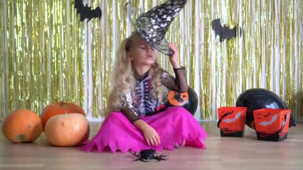 Klein boos blond meisje met zet op heks hoed en kijk naar de camera in de buurt pompoenen - Video