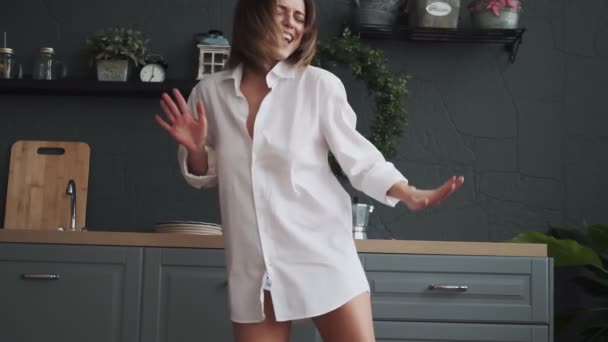 young woman funny and carefree dancing in the kitchen in a white shirt. happy girl having fun and carefree dancing at home in the early morning - Video