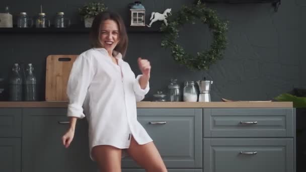 happy girl rejoices and carefree dances in the kitchen. young woman in a white mans shirt carefree dancing at home - Кадри, відео