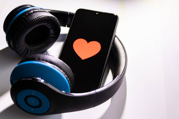 Czarny smartfon z czerwonym sercem na czarnym ekranie i bezprzewodowym niebieskim przez słuchawki słuchowe show mobile music streaming, kontrola zdrowia, uzależnienie od smartfonów, miłość do muzyki i słuchanie muzyki - Zdjęcie, obraz