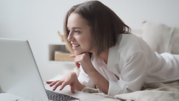 young woman uses a laptop while lying in bed. girl working on a laptop in the morning, comfortably sitting in bed - Séquence, vidéo
