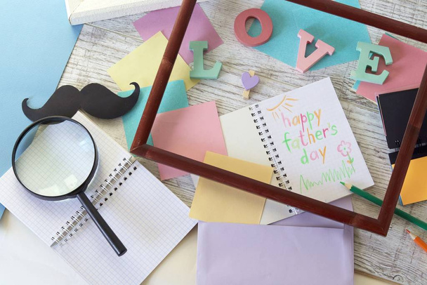 Preparation for the celebration of Father's Day, a composition of three-dimensional letters, frames, funny mustaches, magnifying glasses, notebooks, greeting lettering, paper, pencils on the table, top view  - Фото, зображення