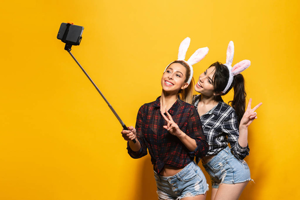 Two Young sexy caucasian woman wearing cute easter rabbit ears making selfie using stick over yellow isolated background - Foto, immagini