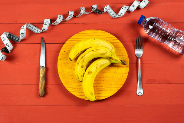 Bananas maduras em uma placa de madeira com talheres em uma mesa de madeira vermelha
 - Foto, Imagem