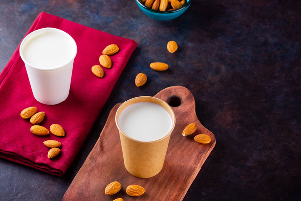 Almond milk with almond on dark background. Homemade organic almond milk in a paper cup on a wooden board. Vegan milk from almonds nuts in kraft paper cup. Alternative milk. Zero waste - 写真・画像