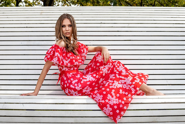 Beautiful young girl, model, brown-haired woman with long hair in a red dress sits on a bench and poses with a relaxed, languid look. - Fotografie, Obrázek