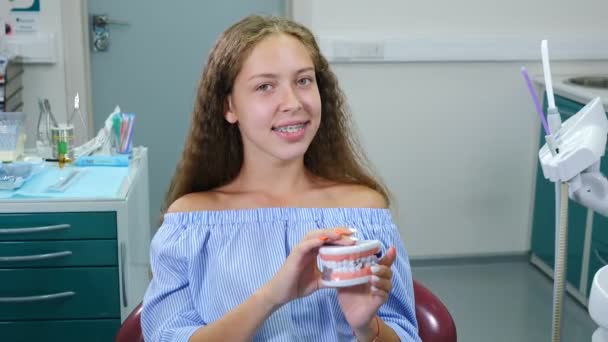 Glimlachend meisje met orthopedische tandbeugels in een tandartsstoel. Gezondheidszorg, tandheelkundige gezondheid en tandheelkunde concept. gelukkig jong Kaukasisch meisje Kijken naar camera poseren op tandheelkundig kantoor achtergrond voor video - Video