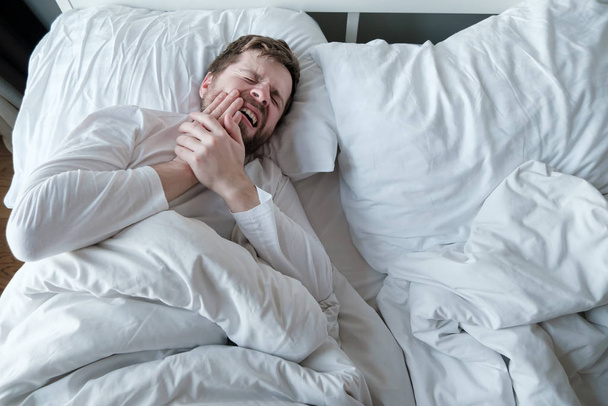 Mies valkoisessa pyjamassa kärsii vakavasta hammassärystä, hän pitää kädestä poskellaan, makaa sängyssä, talossa, luonnonvalossa.
. - Valokuva, kuva