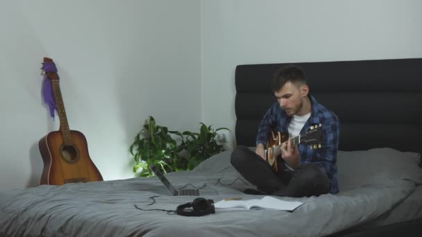 Man creating new song on electric guitar. Musician is taking notes and practicing guitar lessons. Young millennial is learning guitar solo sitting on bed at living room. Music concept - Materiał filmowy, wideo