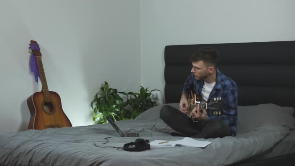 Man plays rock on guitar. Young musician playing on electric guitar sitting on bed at modern living room. Male practicing guitar. Emotional millennial performing his new song. Music concept - Imágenes, Vídeo