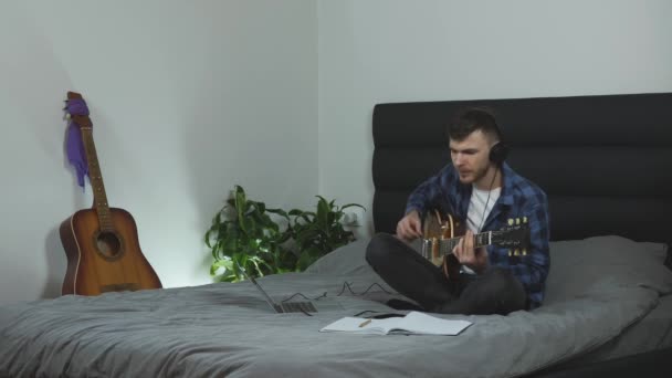 Guitar musician plays on guitar and sings a song. Young man in headphones creating song and taking notes. Caucasian guy learning lyric music on electric guitar. Guitar lessons. Music concept - Metraje, vídeo
