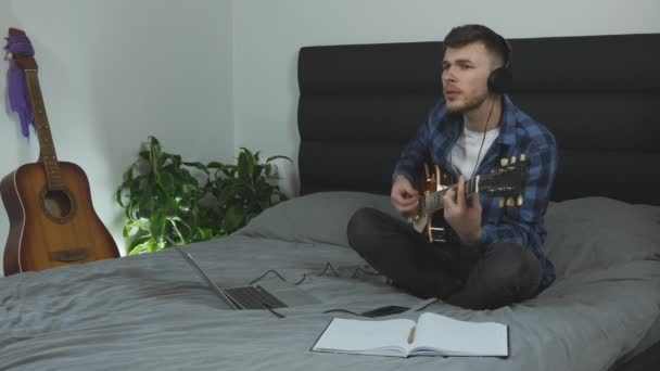 Male emotionally singing and playing on electric guitar. Young handsome man plays on guitar at home. Musician learning new song. Guy in headphones performs lyric music on bed in modern living room - Metraje, vídeo