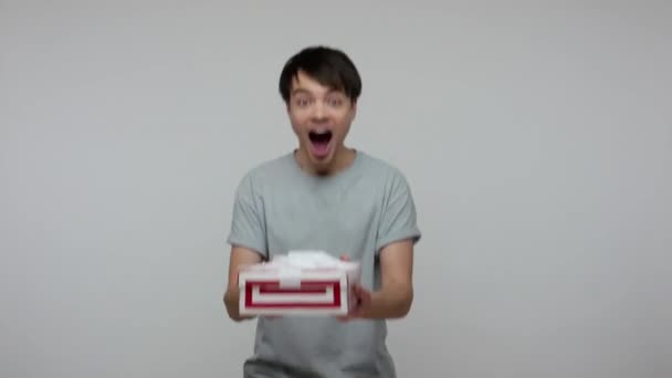 Happy lucky smiling guy in casual t-shirt catching wrapped gift, shaking and embracing box, satisfied with good present, bonus or birthday surprise. indoor studio shot isolated on gray background - Footage, Video