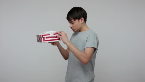 Unlucky brunette man in T-shirt opening long-awaited present box and looking with disappointed upset expression, dissatisfied with birthday gift. indoor studio shot isolated on gray background - Footage, Video
