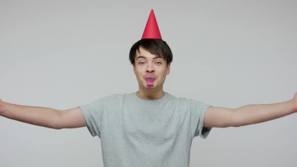 Overjoyed happy man with funny cone on head blowing party horn and dancing under falling glitter confetti, celebrating birthday or success in career. indoor studio shot isolated on gray background - Кадры, видео
