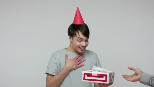 Joyful smiling young guy with funny cone on head receiving present, opening box with happy amazed expression, pleasantly surprised by birthday gift. indoor studio shot isolated on gray background - Imágenes, Vídeo
