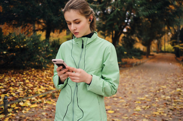 Hoş bir sonbahar parkında cep telefonu kullanan kulaklıklı çekici bir kız. - Fotoğraf, Görsel