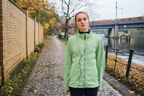 Attractive sporty girl listening music in earphones after run in autumn city park - Фото, зображення