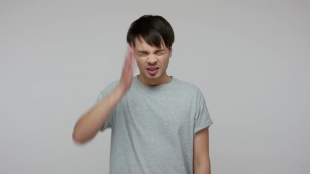 Upset sorrowful young man in T-shirt gesturing facepalm, holding hand on forehead expressing desperate emotions, regretting deeds, accusing himself. indoor studio shot isolated on gray background - Filmagem, Vídeo