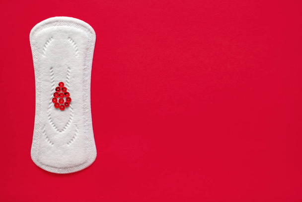 Almohadilla menstrual con destellos rojos en forma de una gota de sangre sobre un fondo rojo, vista superior, miniatura del período menstrual
 - Foto, Imagen