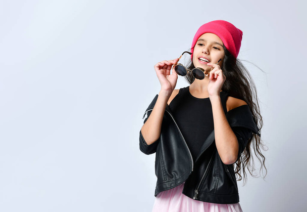 Teenage child in black leather jacket, skirt and pink hat. She is holding her sunglasses, posing isolated on white. Close up - Φωτογραφία, εικόνα