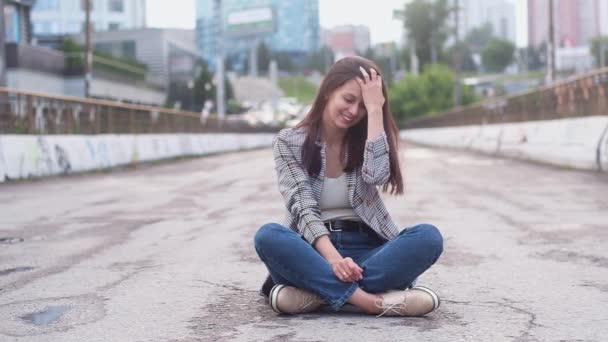 Porträt eines erwachsenen schönen Mädchens mit langen schwarzen Haaren. Eine Frau sitzt auf einer Brücke auf der Straße im Hintergrund der Stadt. Er glättet seine Haare und lächelt in die Kamera. Hipster. Unabhängige Frau. - Filmmaterial, Video
