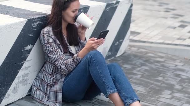 volwassen meisje in een casual jasje en jeans zit op de stoep in de buurt van het beton verdelen gestreepte blok en drinkt warme koffie of thee uit een papieren Beker. Hipster gebruikt de telefoon en leest het bericht. close-up - Video