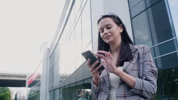 business woman in casual clothes with a modern phone in her hands stands on the background of the business center. A beautiful girl flips through the Internet or messages on her smartphone and smiles. - Filmati, video