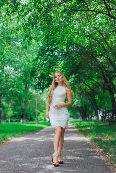 Portrait of a charming blond woman wearing beautiful white dress standing on the road under trees. - Foto, immagini