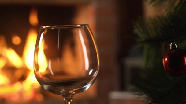 transparent empty wine glass stands near a small Christmas tree with decorations on the background of a burning fireplace. Christmas. Red new-year balls. Celebration and fun by the fireplace. - Imágenes, Vídeo