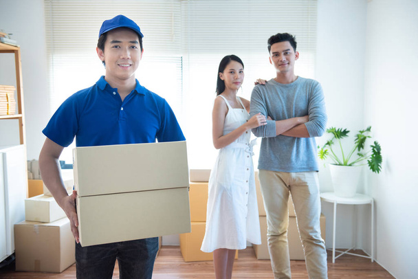 Sorrindo jovem asiático entrega homem em azul uniforme segurando e carregando dois cardbox pacote olhar para a câmera com feliz jovem asiático casal remetente de pé no fundo
. - Foto, Imagem