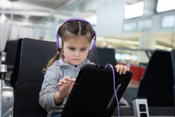 Kulaklıklı küçük kız havaalanında tabletle oynuyor. - Fotoğraf, Görsel