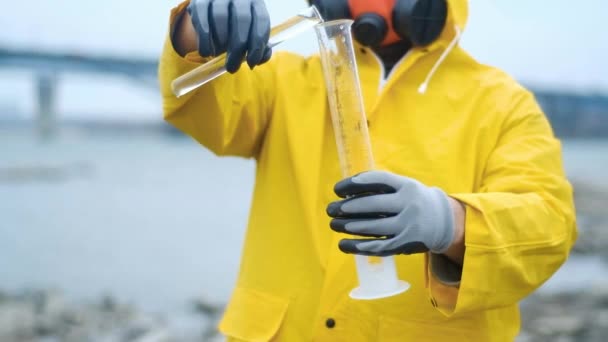biotech expert in a respirator pours water from the river into a test tube for bacteriological analysis. Carefully examines the contents. Shakes to determine contamination. Ecology is modern. - Footage, Video
