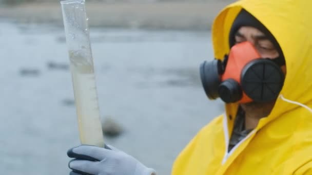 Ein Atemschutzgeräteträger gießt Wasser aus dem Fluss zur bakteriologischen Analyse in ein Reagenzglas. Untersucht sorgfältig den Inhalt. Schüttelt auf, um die Verschmutzung zu identifizieren. Moderne Ökologie. - Filmmaterial, Video