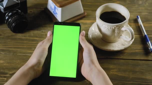 girl is sitting at home at wooden table and holding smartphone with a green screen. teen gesture simulates scrolling up messages or the Internet or pictures on the screen with your finger. Chromakey - Footage, Video