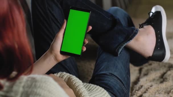 dívčí gesto listuje fotografiemi prsty pomocí svého zeleného displeje telefonu. smartphone chromakey. žena odpočívá na gauči a drží smartphone. Použijte zelený mockup pro vložení fotografie nebo obrázku. - Záběry, video