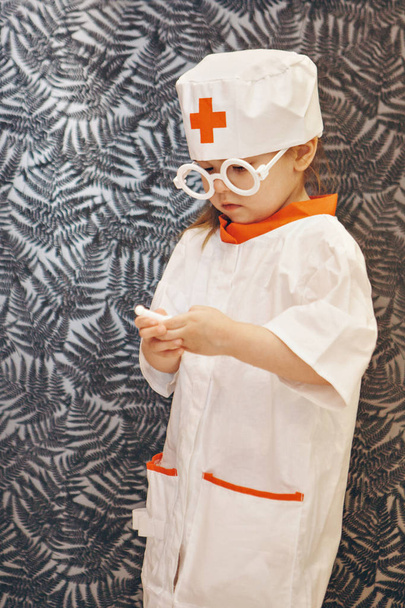 a little girl in a doctor's suit. The child is playing doctor. White coat, glasses, cap with a red cross, medical equipment. Concept of a healthy lifestyle, health care, medicine - Zdjęcie, obraz