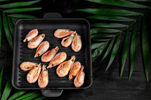 Grilled prawns in a pan with a sprig of rosemary for frying. On the substrate are green palm leaves. On a black wooden background. - 写真・画像