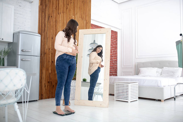 Young woman in a beige blouse standing on a weigher and looking at the mirror - Foto, Imagen
