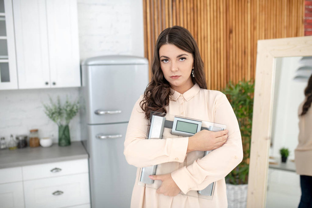 Young woman in a beige blouse standing with weigher and looking upset - Photo, Image