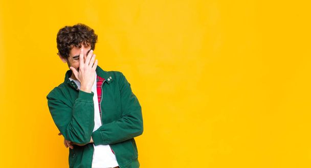young crazy bearded man looking stressed, ashamed or upset, with a headache, covering face with hand against flat wall - 写真・画像