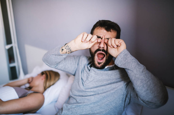 Giovane uomo barbuto sbadigliando, strofinando gli occhi e cercando di svegliarsi presto la mattina mentre sua moglie dorme
. - Foto, immagini