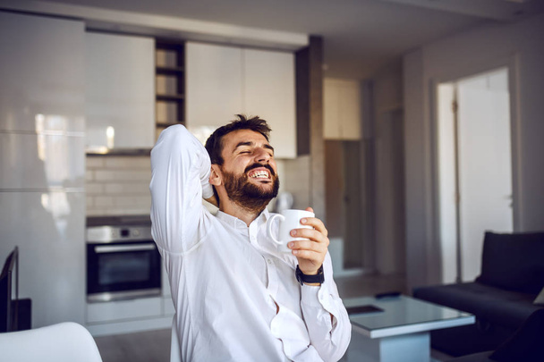 Νεαρός χαμογελαστός ελκυστικός γενειοφόρος άνδρας κάθεται στο σπίτι κρατώντας κούπα με φρέσκο πρωινό καφέ και χαλάρωση. - Φωτογραφία, εικόνα