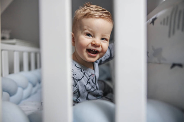Adorable laughing blonde little baby boy with beautiful blue eyes sitting in his crib in the morning and looking at his parent. - Fotoğraf, Görsel