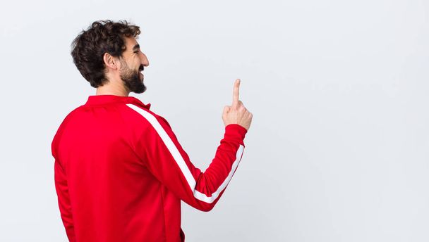 young bearded man back view smiling cheerfully and happily, pointing upwards with one hand to copy space against copy space wall - Fotoğraf, Görsel