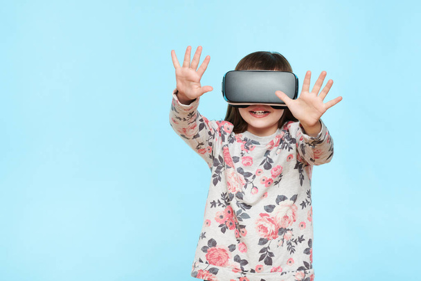 Studio portrait of cute young caucasian girl wearing virtual reality glasses against pastel blue background. Future gadgets technology concept. - Photo, Image