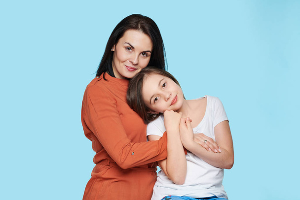 Waist up studio portrait of young mother embracing her daughter. Happy smiling family isolated over pastel blue background. - 写真・画像