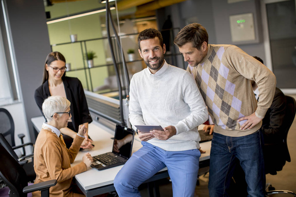 Grupo de empresarios que trabajan y se comunican mientras están de pie en la oficina junto con sus colegas sentados en segundo plano
 - Foto, imagen